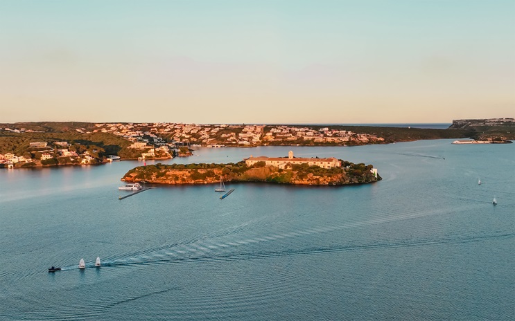 An island with historic buildings is surrounded by calm blue water and small boats at sunset.