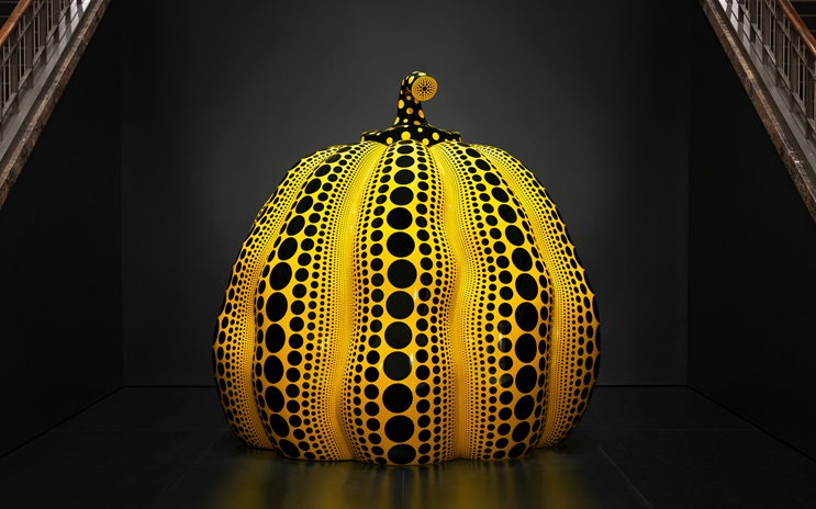 A large yellow pumpkin sculpture covered with black dots sits in a dark room.