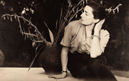 Jacqueline Roque sits on the ground, leaning to one side with her hand on her head, near a large plant.