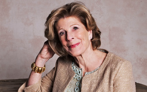 An older woman with styled hair and a gold jacket is smiling and resting her head on her hand.