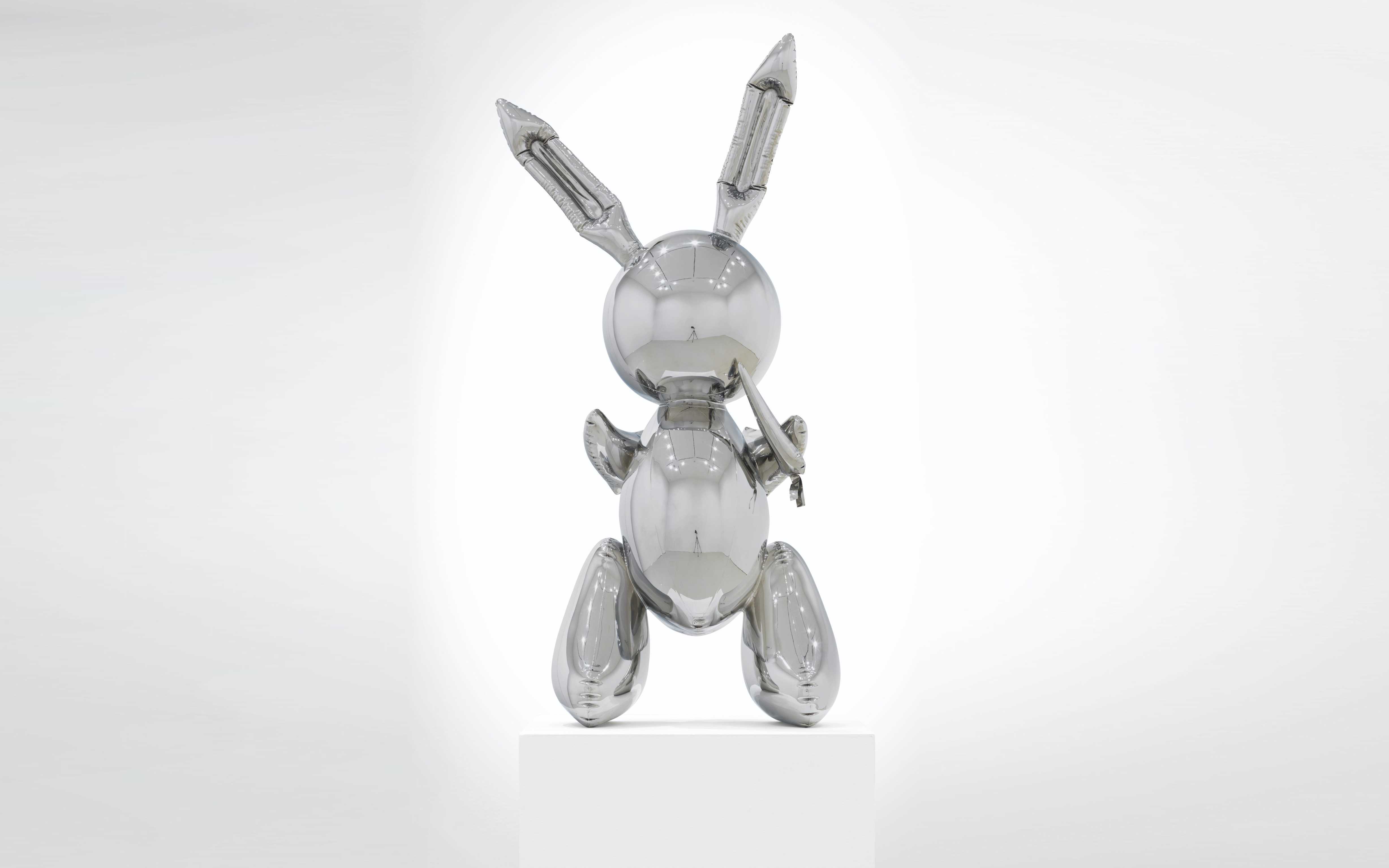 A reflective, metallic sculpture of a rabbit, shown in a sitting pose on a white pedestal against a gradient background.