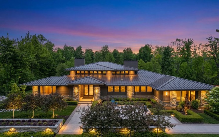 A modern house with a metal roof is illuminated at dusk, surrounded by trees and landscaped gardens.