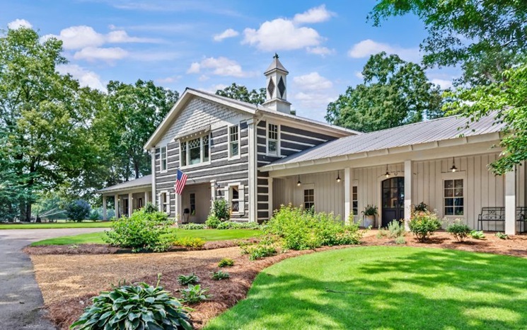 A rustic house with gray siding, a cupola, landscaped gardens, and an American flag.