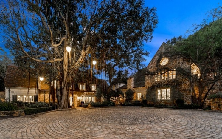 A cobblestone driveway leads to a large stone house surrounded by trees and glowing lights at dusk.