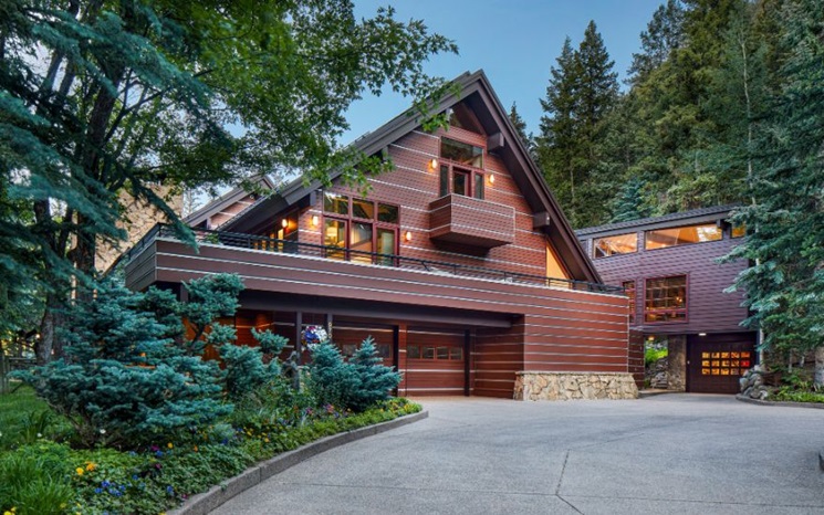 A modern brown house with large windows, surrounded by trees and landscaped greenery.