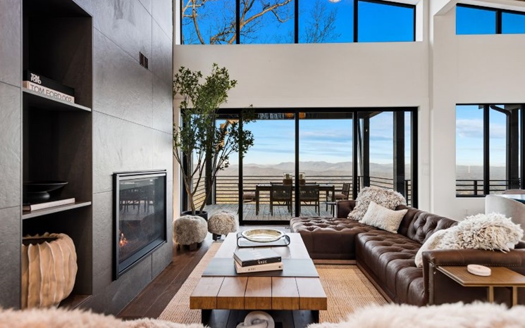 A modern living room with a large leather sectional, fireplace, and floor-to-ceiling windows.