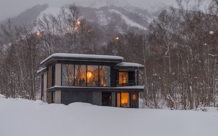 A modern house with large windows glows warmly amid a snowy forest and mountain landscape.