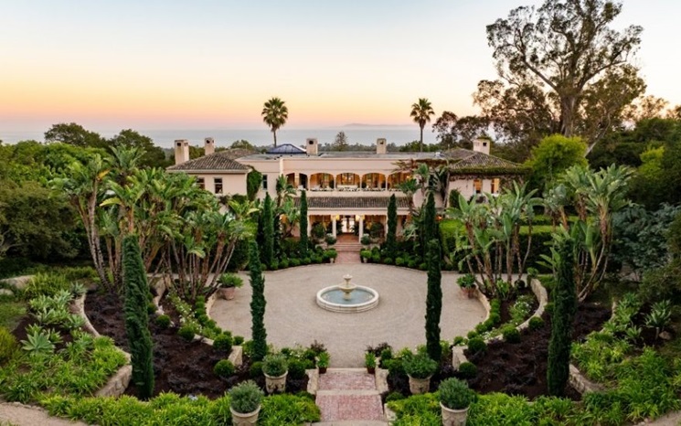 A luxurious mansion with a central circular fountain, lush gardens, and palm trees is shown at sunset with ocean views in the background.