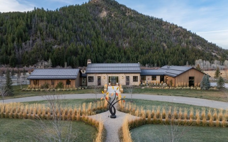 A modern house with a metal roof sits in front of a forested mountain and a large sculpture.