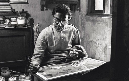 A person is painting at a table in a dimly lit room with art supplies nearby.