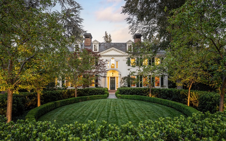 A grand white mansion with manicured lawn and hedges, framed by trees and evening lighting.
