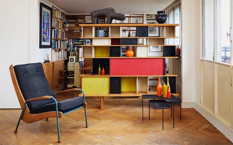 A modern living room features a black lounge chair, colorful shelving unit, vibrant vases, and parquet wooden flooring.