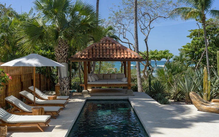 A tropical outdoor pool area with lounge chairs, an umbrella, and a shaded gazebo near the ocean.