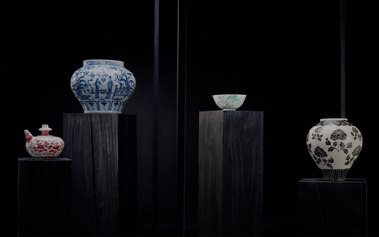 Four decorative ceramic pieces are displayed on dark wooden pedestals against a black background.