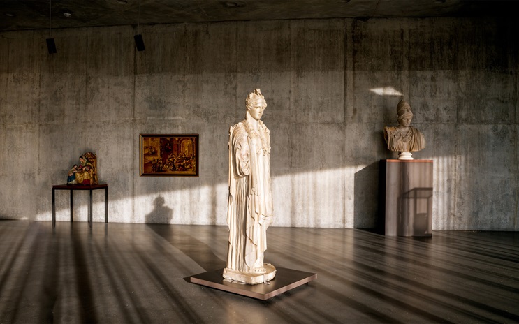 A gallery with classical statues, a painting, and a sculpture on display against concrete walls.