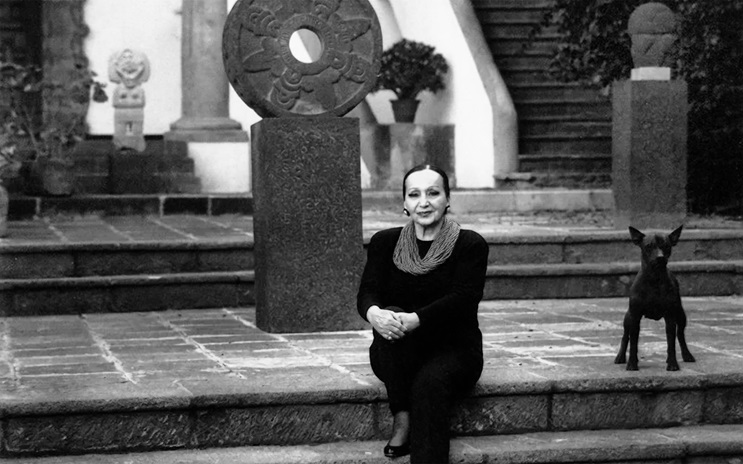 Dolores Olmedo wearing black sits on stone steps beside a small dog, surrounded by sculptures and plants.