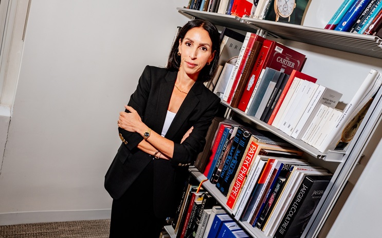 Rebecca Ross in a black suit stands with folded arms next to a bookshelf filled with books.