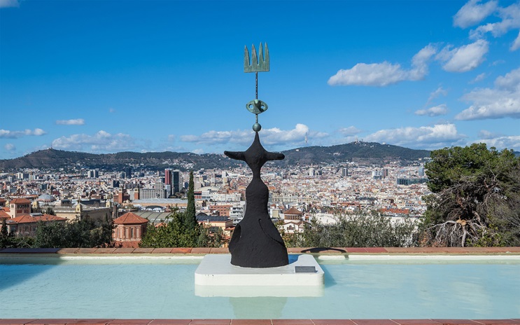 A modern sculpture stands in a shallow pool overlooking a cityscape and distant hills.