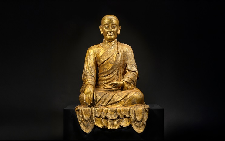 A golden statue of a seated figure in traditional robes against a dark background.