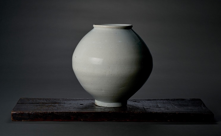 A round, white ceramic vase sits on a dark wooden surface against a plain background.