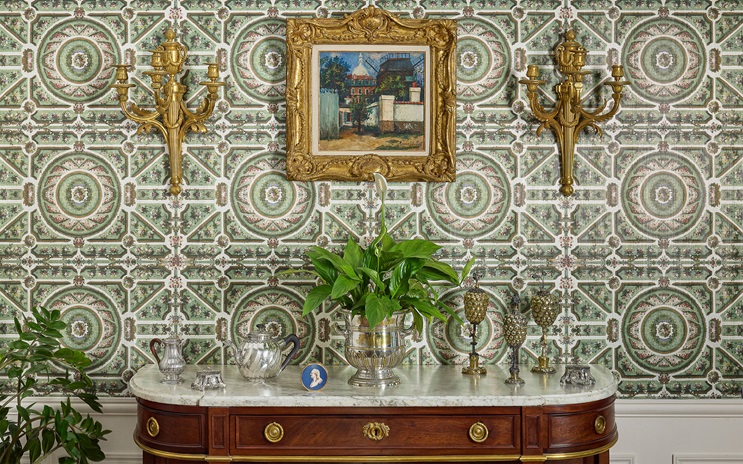 A vintage sideboard with ornate decor sits against patterned wallpaper and a framed painting.