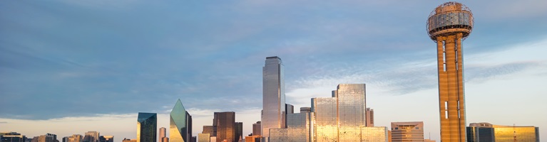 A city skyline with modern skyscrapers and a tall tower under a partly cloudy sky at sunset.