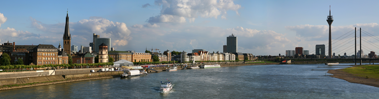 Panoramic view of a river flowing through Düsseldorf with buildings, a church, a tower, and a modern suspension bridge.