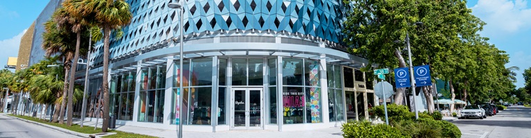 A modern glass building with a geometric facade stands on a sunny, palm-lined street corner.