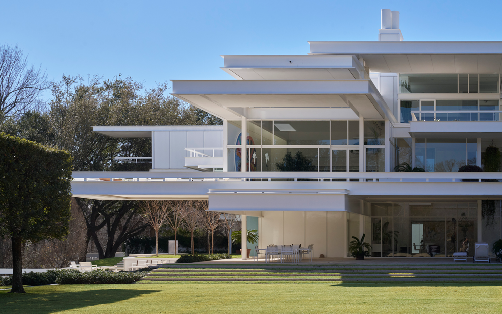 A modern white house with large glass windows and wide horizontal overhangs sits beside a green lawn
