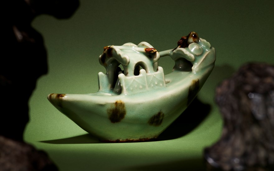 A close-up of a celadon incense burner shaped like a mythical creature, shadowed by rocks, on a green background. | A ceramic piece resembling a boat with intricate details and discoloration, set against a green background.