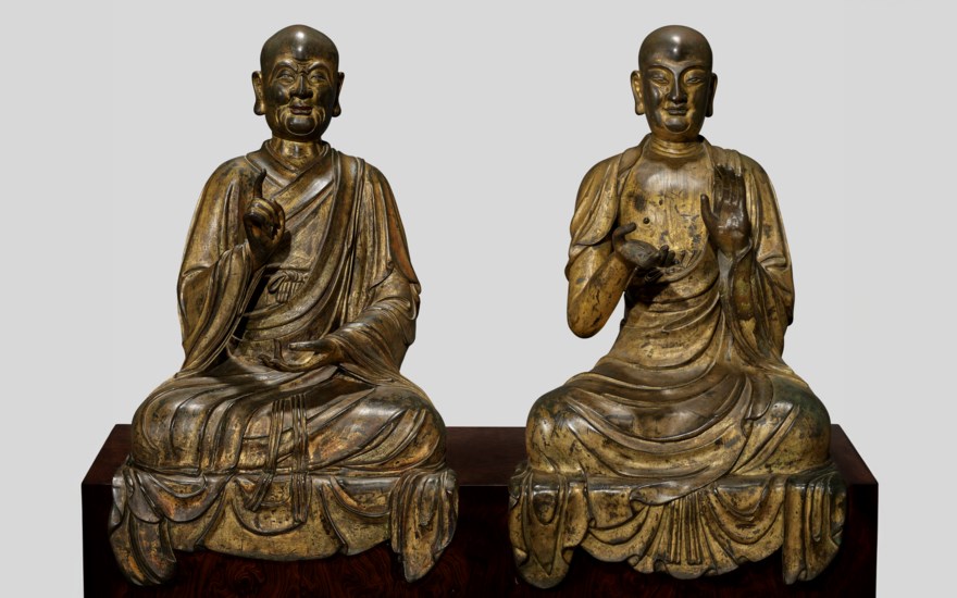 Two golden statues of seated figures in robes with blurred faces, displayed on a wooden platform.
