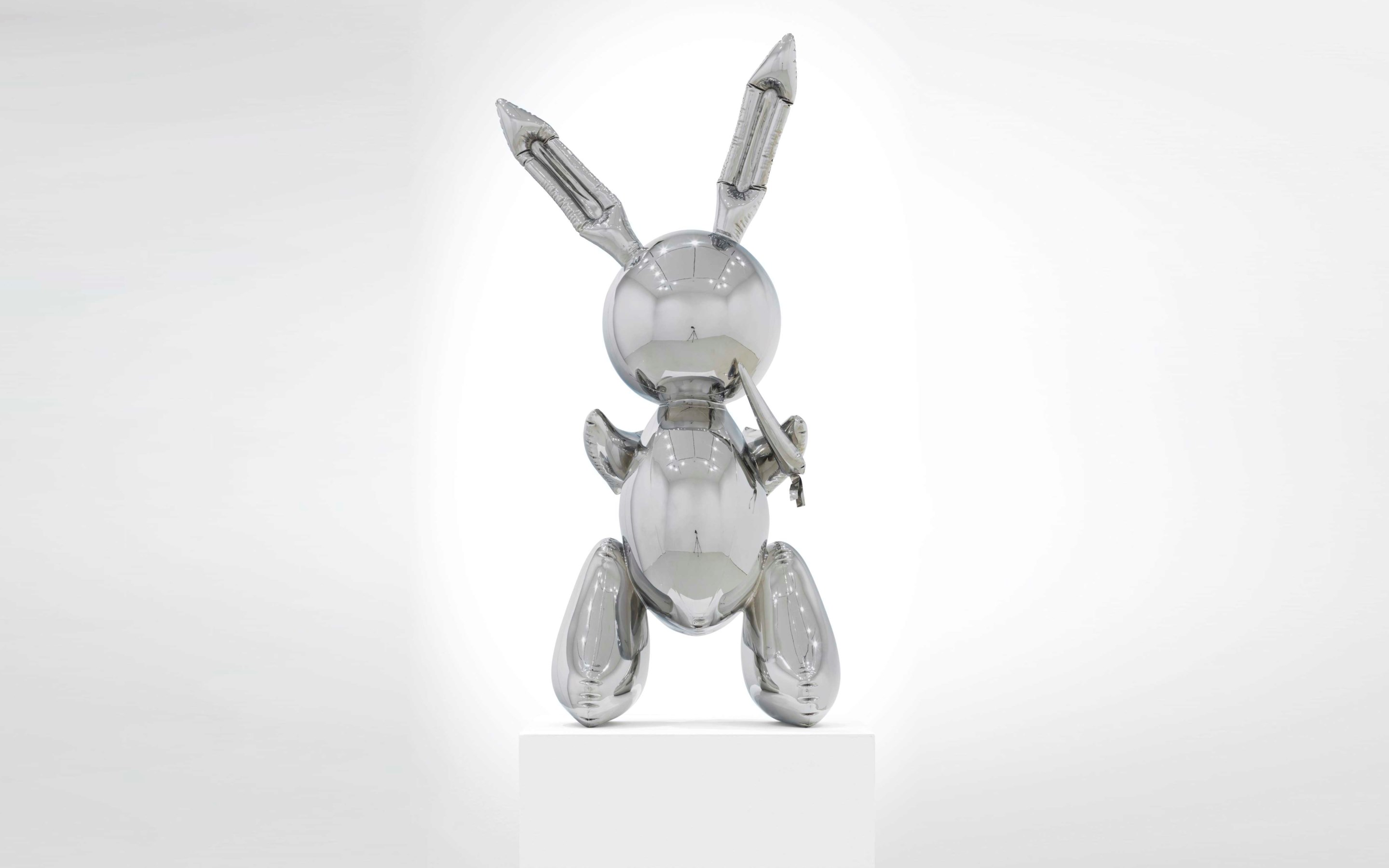 A shiny, metallic rabbit sculpture stands upright on a white pedestal against a plain background.