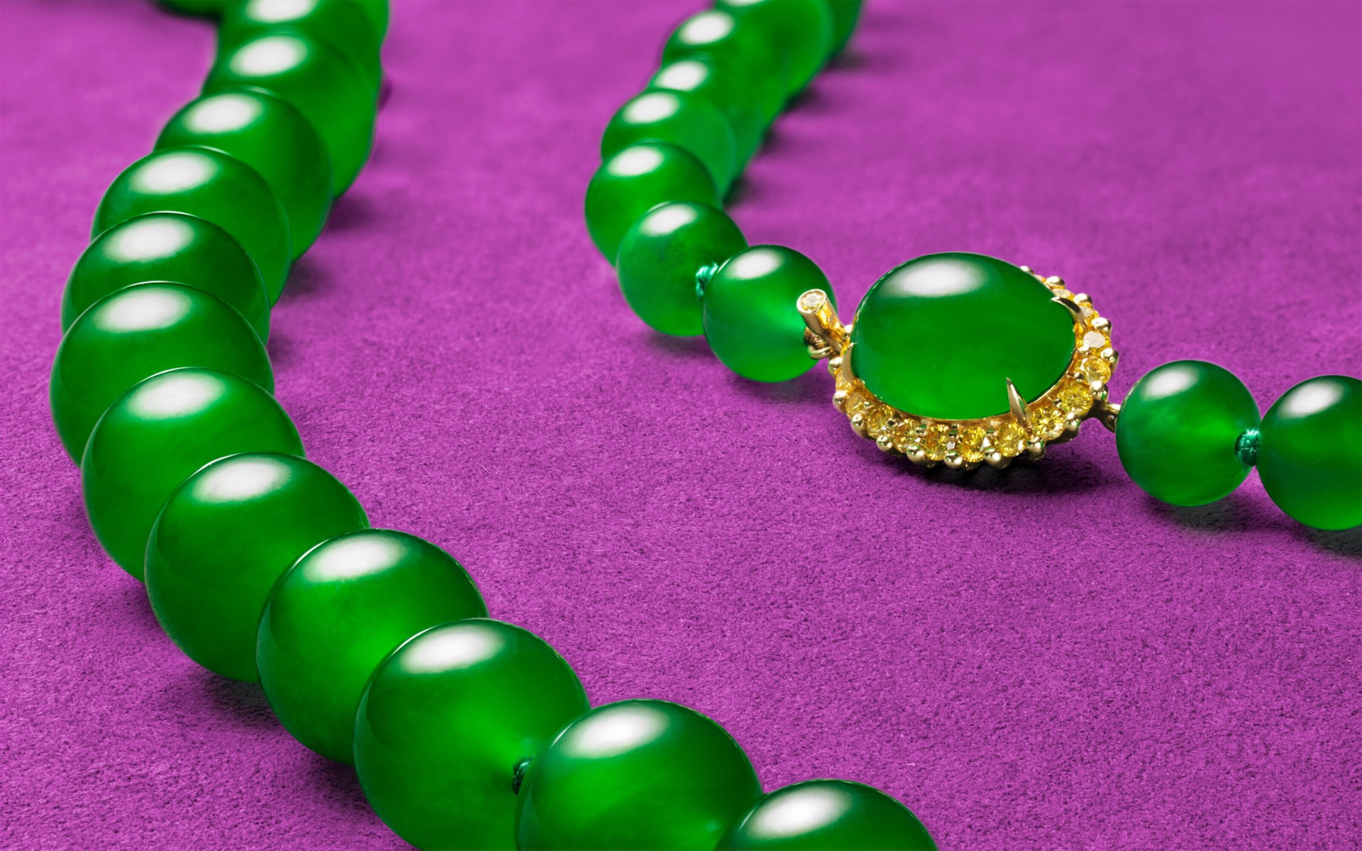 A green jade beaded necklace with a gold clasp is displayed on a purple fabric surface.