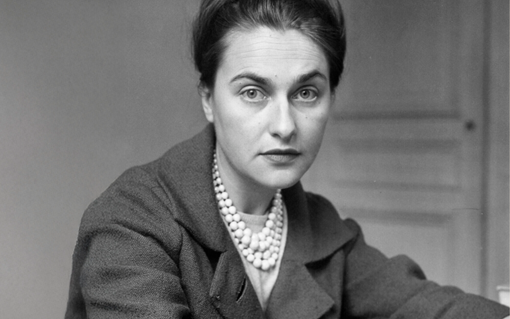A woman with dark hair in an updo, wearing a pearl necklace and coat, looks at the camera.
