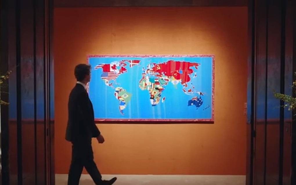 A man in a suit walks past a colorful world map painting displayed on an orange wall.