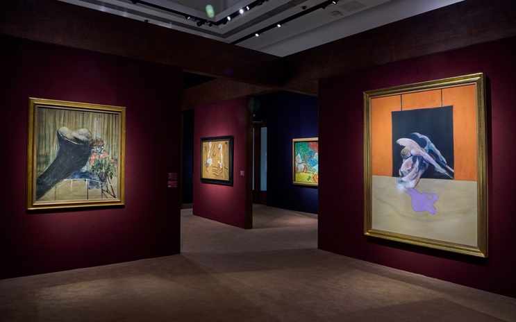 A dimly lit art gallery displays several modern paintings in gold frames on deep red walls.