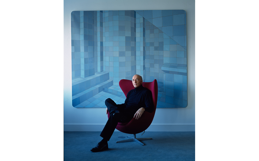 A man in black sits on a red chair in front of a large blue geometric painting.