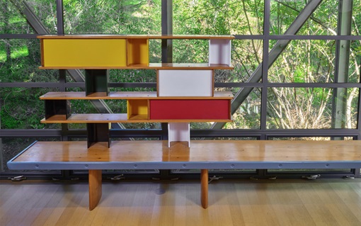 A colorful, modern shelving unit sits on a wooden bench in front of large glass windows.