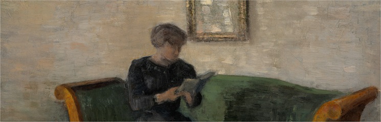A person in dark clothing sits on a green sofa reading a book in a softly lit room.