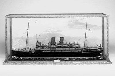 A detailed waterline sailor's model of H.M.S. Macedonia, (1914-1918) 
with masts, yards, standing rigging, radio aerial, anchors, wood capped deck rails, fairleads, bollards, capstan, bollards, winches, ventilators, hatches, companionways, cranes with rigging, superstructure with glazed bridge with open bridge over with binnacle, helm, telegraph, searchlights, deck lights, two stayed funnels with safety valve extension pipes, engine room lights, ten coverd life boats including one steam launch in davits, deck guns and other details. The hull with portholes, is finished in black and red and mounted on a painted moulded seascape, glazed case with painted sky background and legend -- 22 x 47½in. (56 x 120.5cm.)
See illustration