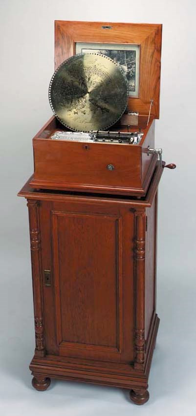 A fine 15¾-inch Symphoniontable disc musical box with side-by-side combs on long bedplate of Art Nouveau design, in walnut case with transfer on lid and monochrome internal print, on an oak pedestal cupboard with two disc storage shelves, containing 175 discs with original lacquered finish -- 55in. (140cm.) high overall. circa 1900-1910