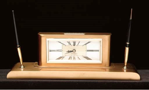 HEFNER, Hugh M. Brass desk clock used by High Hefner in Chicago, 1960s, with two pens at sides, by Seth Thomas, nameplate with initials "H.M.H." mounted at top. Width 16 in. (40.6 cm.)