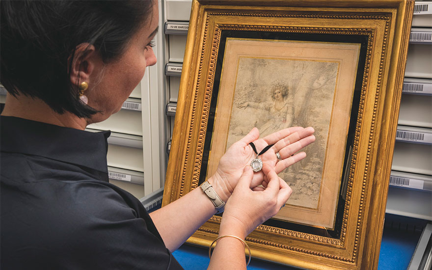 Specialist Jo Langston holding The Maria Fitzherbert Jewel, with a 1789 drawing of Maria Fitzherbert wearing a portrait miniature of the future George IV around her neck, by Richard Cosway R.A.